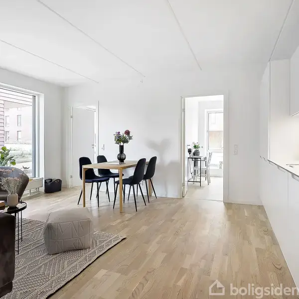 Stue med sorte stole omkring spisebord, sofa ved stor vinduesparti. Moderne køkken integreret i rummet. Lys trægulv og dør fører til andet rum.