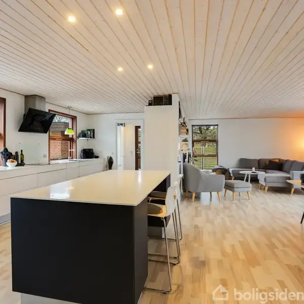 Køkken- og stuerum med moderne, minimalistisk design. Køkkenø med barstole står centralt, omgivet af lyse trægulve. Store vinduer giver udsigt til naturen udenfor. Grå sofa- og spiseområde i baggrunden.