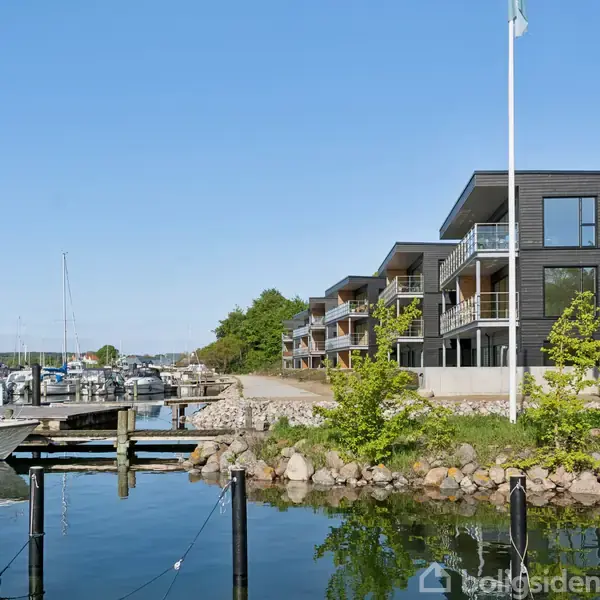 Moderne lejlighedsbygninger ved en marina, spejler sig i det rolige vand. Sejlbåde ligger fortøjet ved bådebroen, mens grønne træer og buske flankerer bygningerne under en klar, blå himmel.