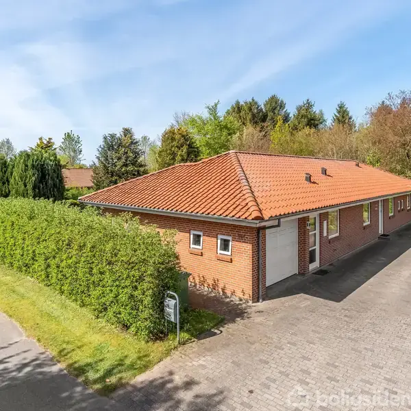 Rødt murstenshus med rødt tegltag står stille ved siden af en nyklippet hæk. En asfalteret indkørsel fører til garagen. Grønne træer omkranser området på en solrig dag.