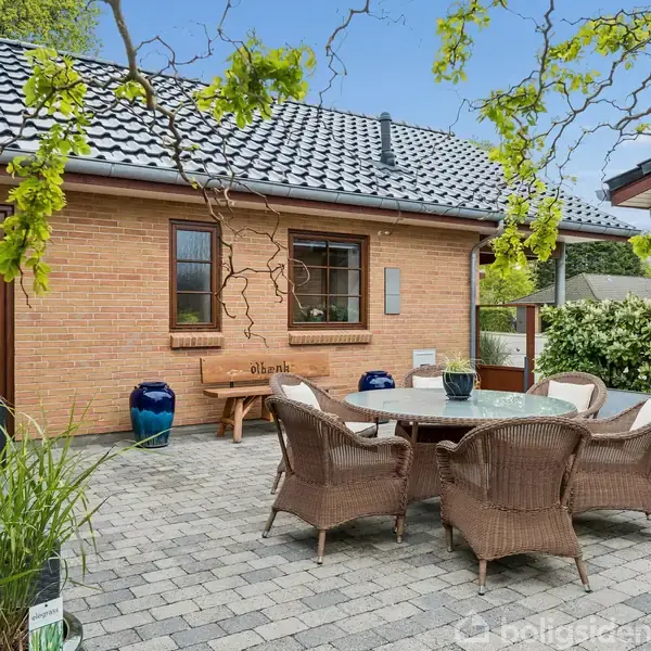 Et rundt havebord omgivet af flettede stole står på en brostensbelagt terrasse. Omkring er der murstensbygninger med sorte tagsten og grønne planter i krukker.