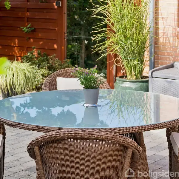 Rundt glasbord med plante i midten står omkranset af fire fletstole på flisebelagt terrasse. Baggrunden viser en murstensvæg, en plante, og et træhegn i solen.