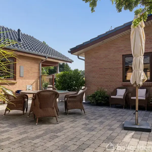 Terrassemøbler står på en flisebelagt patio mellem to murstenshuse. En sammenklappet parasol står ved siden af bordet, omgivet af grønne planter, under en klar blå himmel.