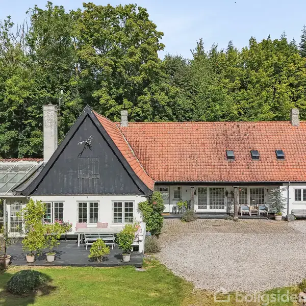 Et hvidt hus med rødt tag står i en skovomgivelse. Terrassen har blomsterkrukker og en parasol. Der er en grusbelagt gårdsplads foran huset.