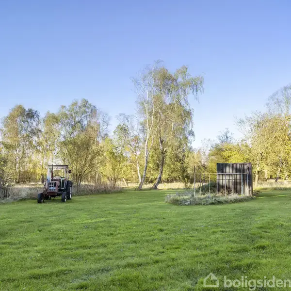 En græsslåmaskine står stille på en grøn græsplæne omgivet af træer i en park eller have.
