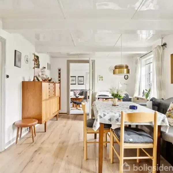 Spisebord dækket med dug, omgivet af stole, står i en lys stue med trægulv. I baggrunden ses en skænk, sofa, og vægbilleder.
