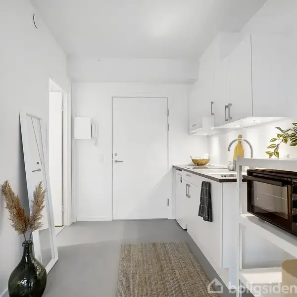 Et hvidt køkken med et komfur og en mikrobølgeovn på en hylde. En grøn plante og en frugtskål står ved vasken. En spejl læner mod væggen ved siden af en vase.