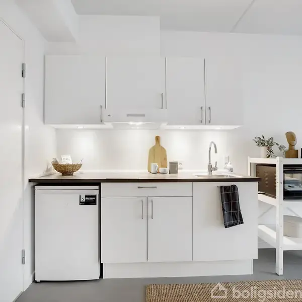 Et kompakt køkken med hvide skabe og en lille håndvask. Et køleskab står under bordpladen. En mikrobølgeovn og kurv er placeret på hylder ved siden af. En dør til venstre.