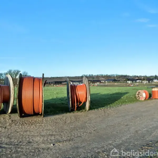 Store træspoler med orange kabler står stille ved en grusvej på en åben mark. I baggrunden ses nogle bygninger og træer under en klar blå himmel.