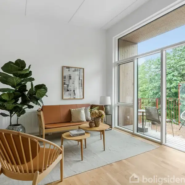 Stue med sofa og stol står på trægulv. Sofaen har puder, og en plante er ved siden af. Store vinduer fører ud til en altan med stole og grønne træer udenfor.