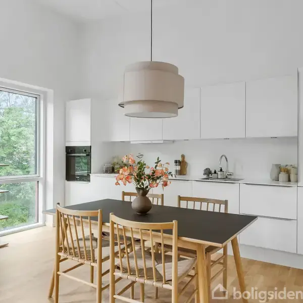 Et moderne køkken med et sort spisebord omgivet af fire træstole. En vase med blomster står på bordet. Hvide skabe og vindue med udsigt til grønne træer.