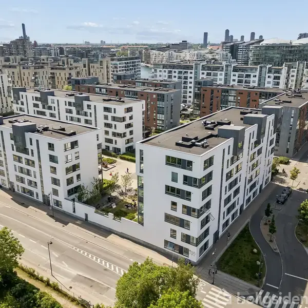 Moderne hvide lejlighedsbygninger står stille i en urban bydel med grønne områder og veje omkring dem, og flere lignende bygninger breder sig ud i horisonten under en klar himmel.