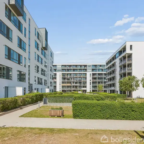 Moderne lejlighedsbygninger står stille omkring en central gårdhave med grønne buske og træer, mens en klar blå himmel strækker sig over det hele.