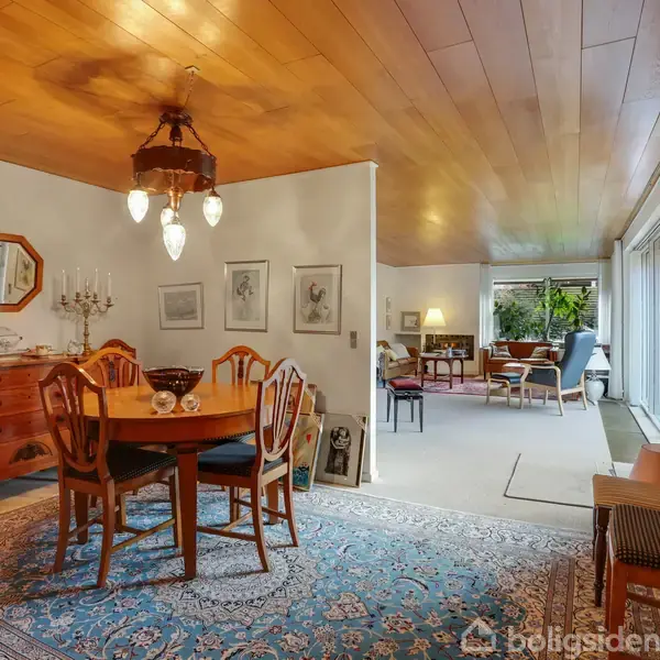 Et spisebord med stole står centralt i et rum. Rundt om er der en kommode, vægkunst og gulvtæppe, i et rum med træloft, lysekrone og store vinduer i baggrunden.