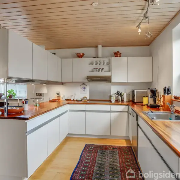 Et hvidt, moderne køkken med trækantede borde, vask og kogezone. Rummet har trægulv, tæppe og vindue til højre. Skabe oppe og nede rummer keramik og køkkenredskaber.