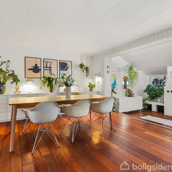 En spisestue med et lyst træbord omgivet af fire hvide stole står på et lakeret trægulv. Planter og kunstværker pryder væggene og det omkringliggende interiør.