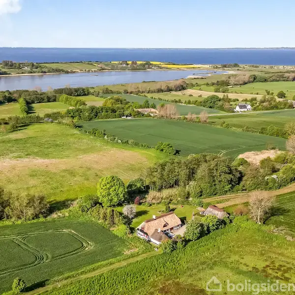 En gård ligger fredeligt omgivet af marker og træer, nær en stor sø i en frodigt, landlig region med udsigt til en fjern horisont ved havet.