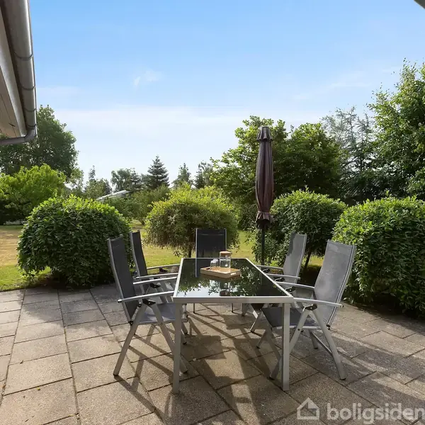 Terrassemøbler står stille på en flisebelagt terrasse. Et langt glasbord er omgivet af fem stole og en lukket parasol. Omkring er der grønne buske og træer under en klar blå himmel.