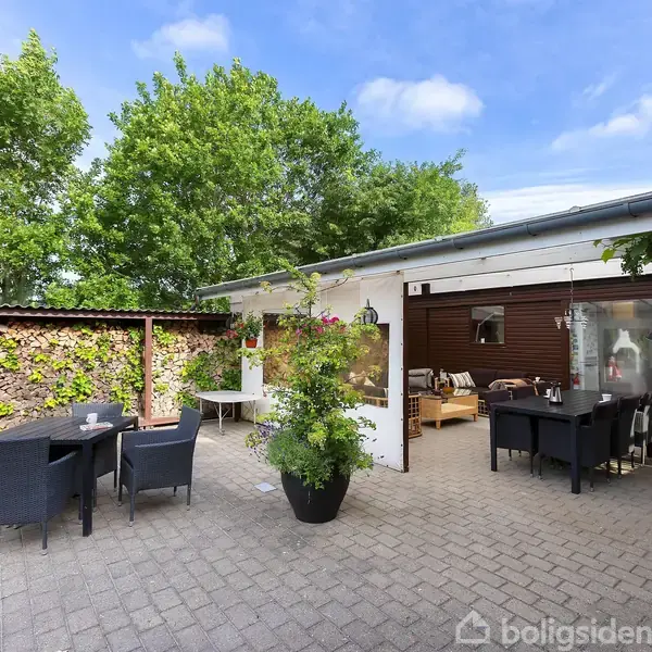 En terrasse med flisebelægning, indeholder et bord og stole på begge sider. I baggrunden er der en pergola dækket af planter. Omgivet af grønne træer og en bygning.