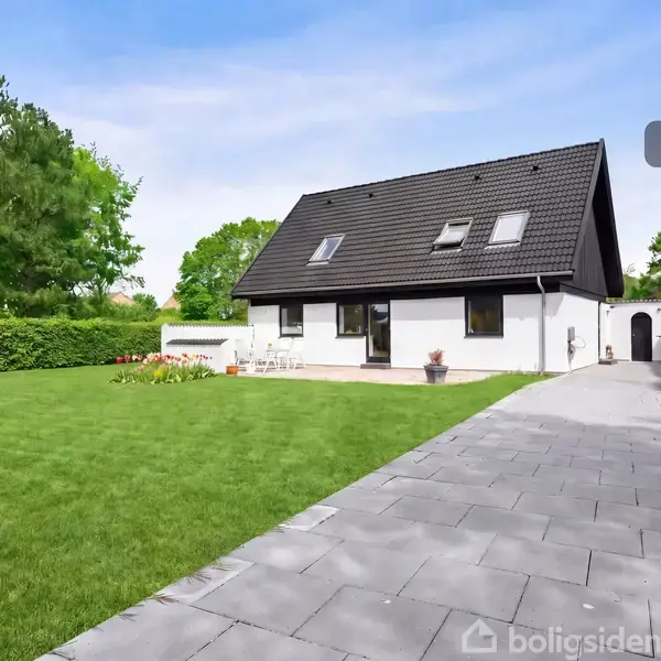 Et hvidt hus med sort tag står i en velplejet have. En flisebelagt sti leder fra forhaven. I baggrunden ses grønne træer og hække. Tekst: "Visualisering".