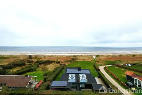 Moderne hus med solpaneler ligger fredeligt ved en kystlinje. Forgrunden viser grønt græs, mens havet strækker sig mod horisonten under en overskyet himmel.