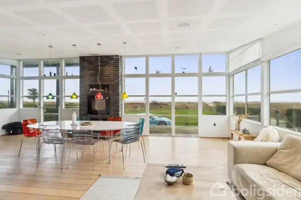 Spisebord med farverige stole står i et lyst rum med panoramavinduer. Udenfor ses en græsplæne og åben natur. En sofa er placeret til højre i billedet.