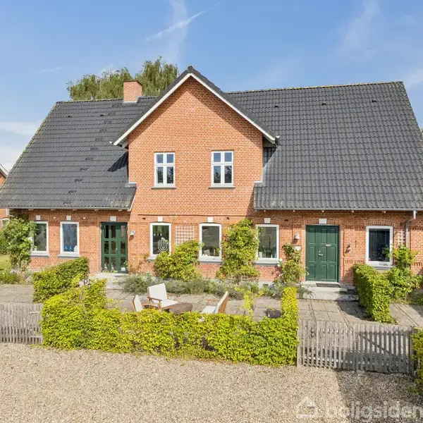 Et rødt murstenshus med to hoveddøre står i en solrig gårdsplads. Grønt løv og små buske dekorerer området. Omridset af nabohuse ses i det grønne boligområde.