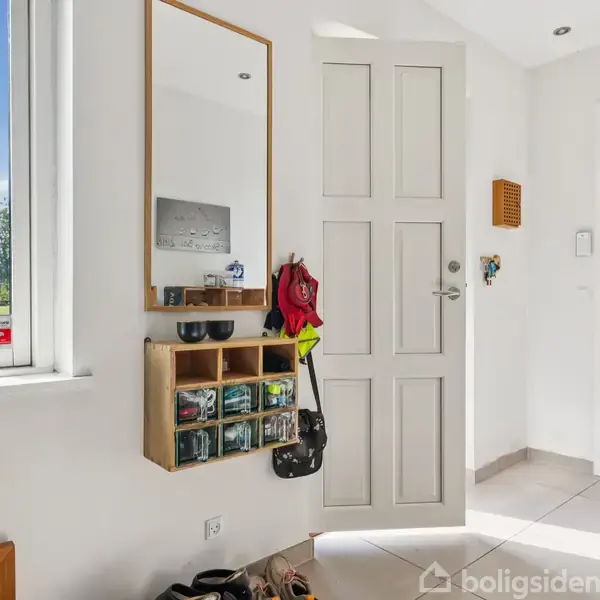 En lys entré med en hvid dør til højre. En vægmonteret hylde indeholder flere opbevaringsbokse og hænger under et spejl. Et vindue viser græs og blå himmel udenfor.