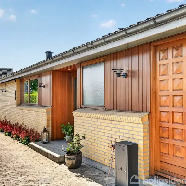 Huset har en trædør, gule mursten og vinduer, flankeret af planter. Det ligger ved en flisebelagt indkørsel i et boligområde med andre huse i baggrunden.