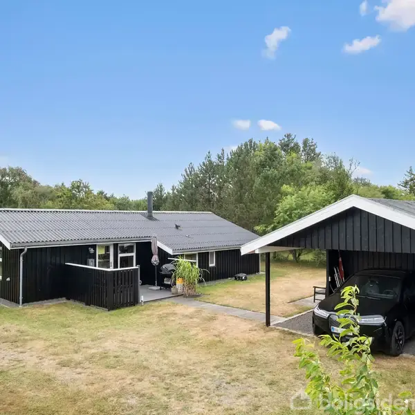 Et sortmalet sommerhus med en tilbygning står på en græsplæne. Ved siden af er der en carport med en bil. Omgivet af træer og blå himmel.
