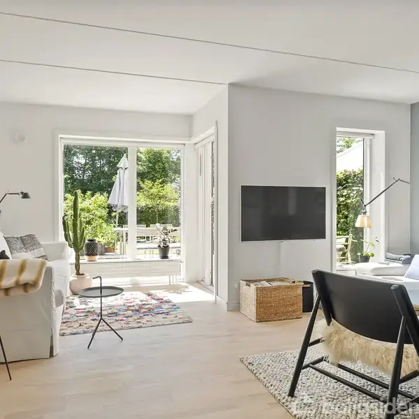 Stue med sofa og lænestol, et sofabord og en gulvlampe. Rummet har store vinduer med udsigt til en terrasse og have. En fladskærm hænger på væggen.