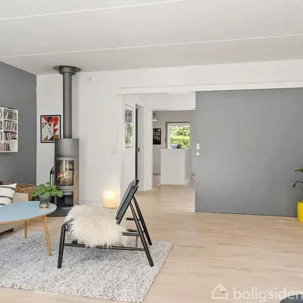 Stue indrettet med sofa og stol omkring et rundt sofabord på et tæppe. En brændeovn i hjørnet. Bogreol til højre, grøn plante i en gul potte. Ingen synlig tekst.