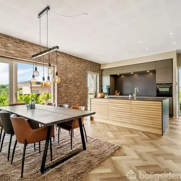 Spisebord med seks stole står på et tæppe, mens moderne lamper hænger over bordet. Køkkenø i baggrunden under et lyst loft. Store vinduer viser en grøn have.