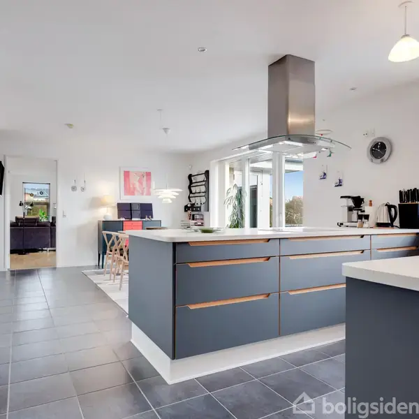 Køkkenø med grå skabe står midt i et moderne køkken. Bordpladen er hvid, omkring er der lysindfald fra vinduer, og et spiseområde ses i baggrunden.