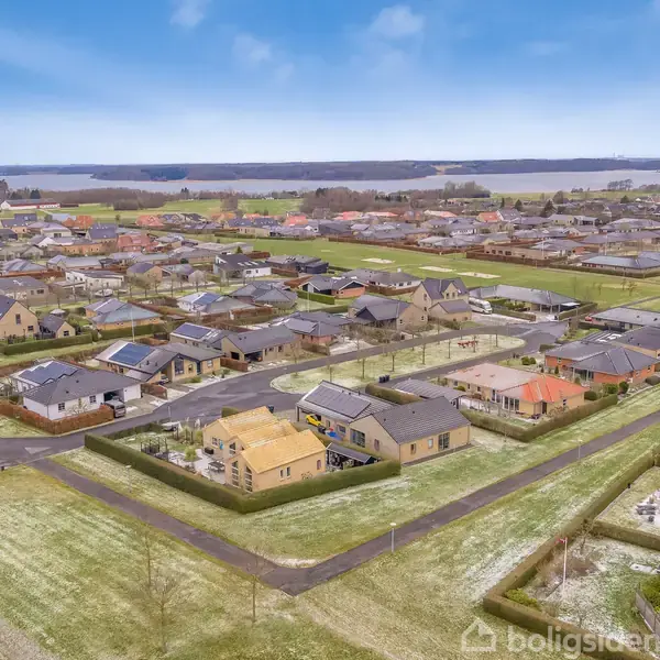 Boligområde med huse omgivet af grønne haver og veje, set fra en fugleperspektiv. Byen ligger nær en sø eller fjord i baggrunden under en klar blå himmel.