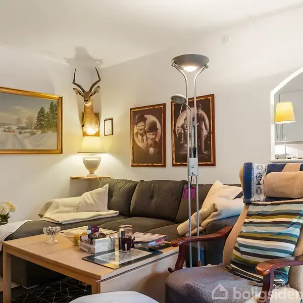 En stue med en hjørnesofa, sofabord og en lænestol med farvet puder. Dekoreret med kunstværker og et dyrehoved på væggen. Indgangen til et køkken ses i baggrunden.