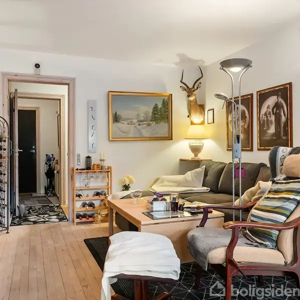 En stue med en lænestol, sofabord og sofa. Væggene prydes af kunst, herunder et hjortehoved. Rummet er oplyst af en gulvlampe og naturligt lys fra køkkenåbningen.
