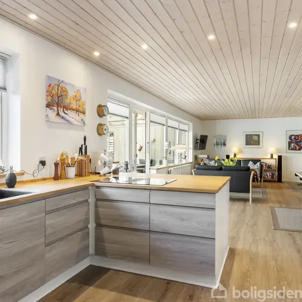 Et moderne køkken med træfinish, hvor kogeplader og køkkenredskaber er placeret på bordpladen. I baggrunden ses en stue med spisebord, sofa og malerier på væggen.