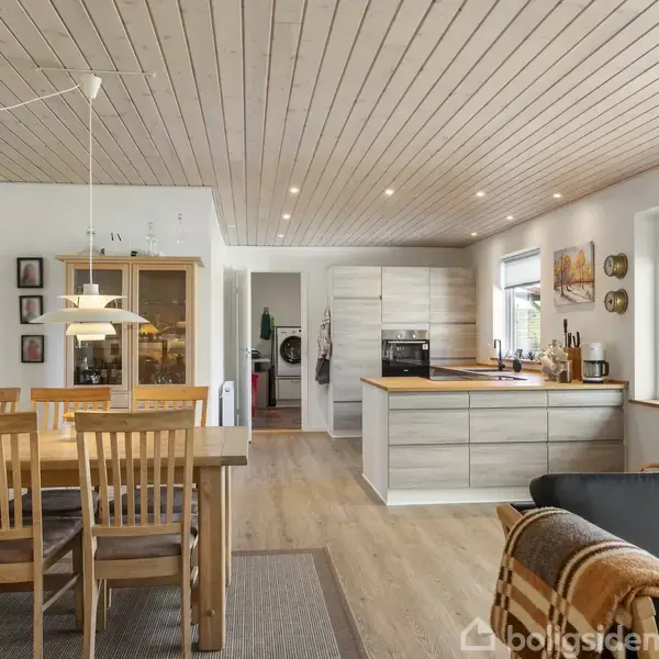 Spisebord med træstole står i en moderne, lyst køkken-alrum. Køkkenet har en stor ø, hvide skabe og indbygningsovn. En sort sofa med et tæppe ligger i forgrunden.