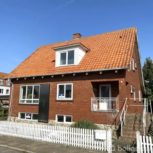 Hus med rødt tegltag og murstensfacade, placeret i en villavej med hvidt stakit rundt om. Sollys rammer taget, mens en trappe leder op til hoveddøren.