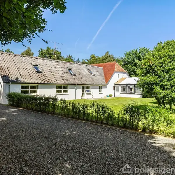 Hus med hvid facade og rødt tag står i sollys, omgivet af træer og grøn have. Grusvej fører op til huset, som har flere vinduer og en terrasse.