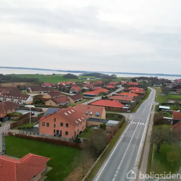 Boliger med røde tage står i rolige rækker langs en vej, der strækker sig gennem et landligt område med græsmarker og en fjord i horisonten under en overskyet himmel.