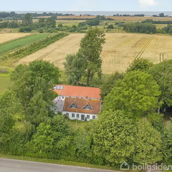 Et hvidt hus med rødt tag ligger omgivet af tæt bevoksning med træer. I baggrunden ses store åbne marker og en blå himmel.