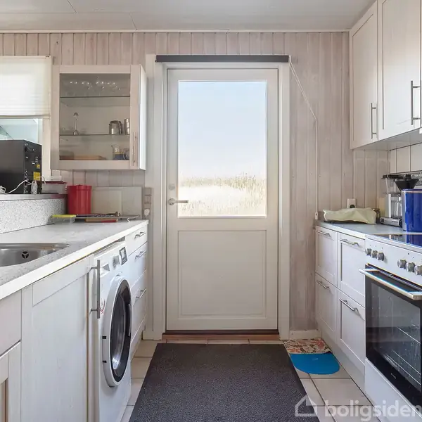 Køkken med hvide skabe og apparater, inklusive vaskemaskine og ovn; lysindfald gennem en glasdør vedrørende en udendørs udsigt. Mat ligger på gulvet foran døren.