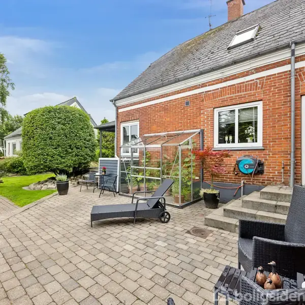 En rød murstenshus har en terrasse med lounge-stole og et lille drivhus. Haven er omgivet af grønne buske og græsplæne, tilbyder en rolig atmosfære.