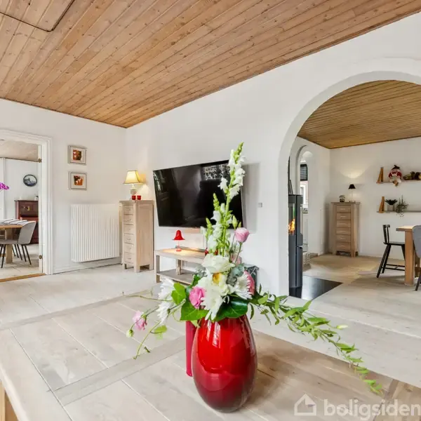 En rød vase med blomster står på et sofabord i en stue med lyst trægulv og loft; indtil et åbent køkkenområde med spisebord og stole ses gennem en bueåbning.