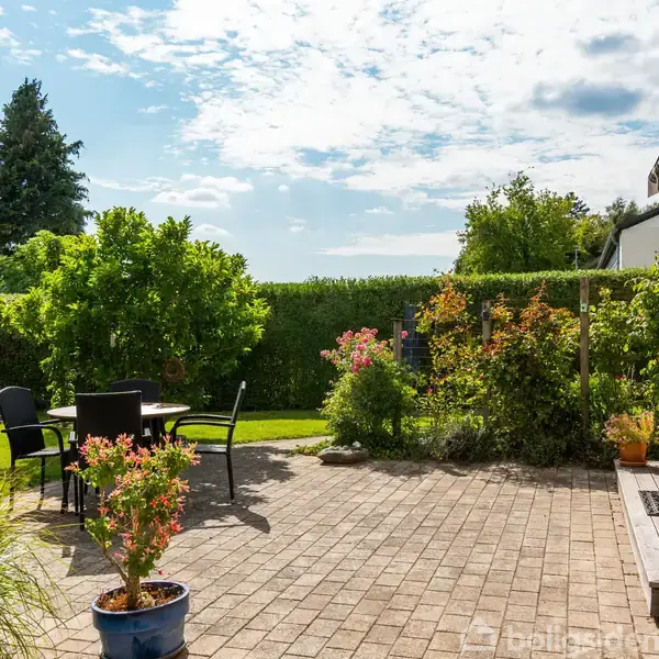 Terrasse med stole og bord står på en brolagt gårdhave. En sort parasol er slået ned. Omgivet af grønne planter og buske, under en klar, let skyet himmel.