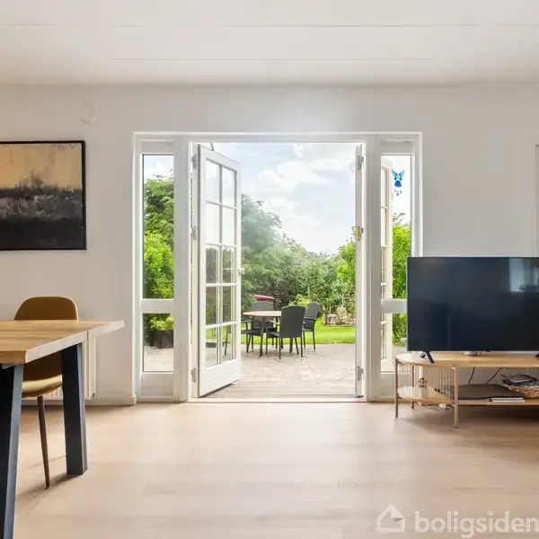 Dobbelt glasdør åbner ud til en terrasse med grøn udsigt. Indendørs: spisebord med stole til venstre, tv på tv-bord til højre, maleri på væggen.