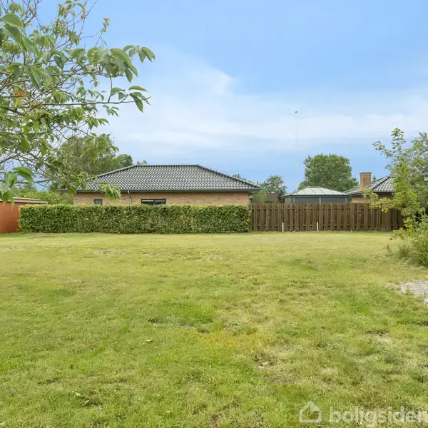 Græsplæne strækker sig bredt ud, omgivet af træer, hække og et hegn, med et rækkehus i baggrunden. En carport med en campingvogn står til venstre.