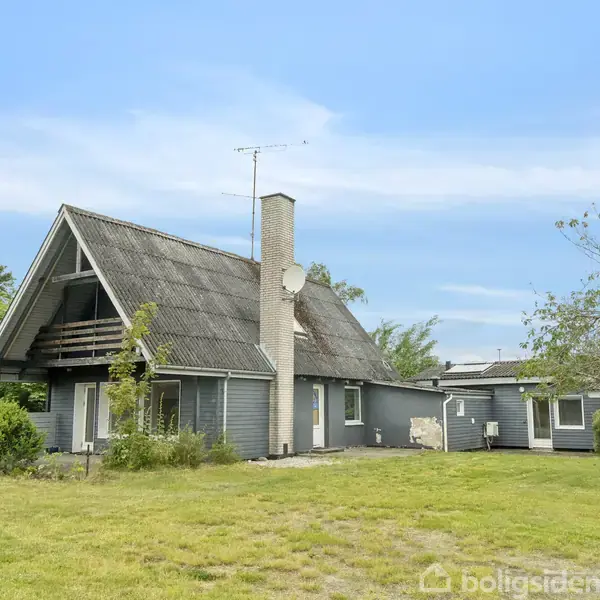 Et gråt, stråtækt hus med skorsten står stille på en velplejet græsplæne omgivet af træer og buske under en blå himmel.